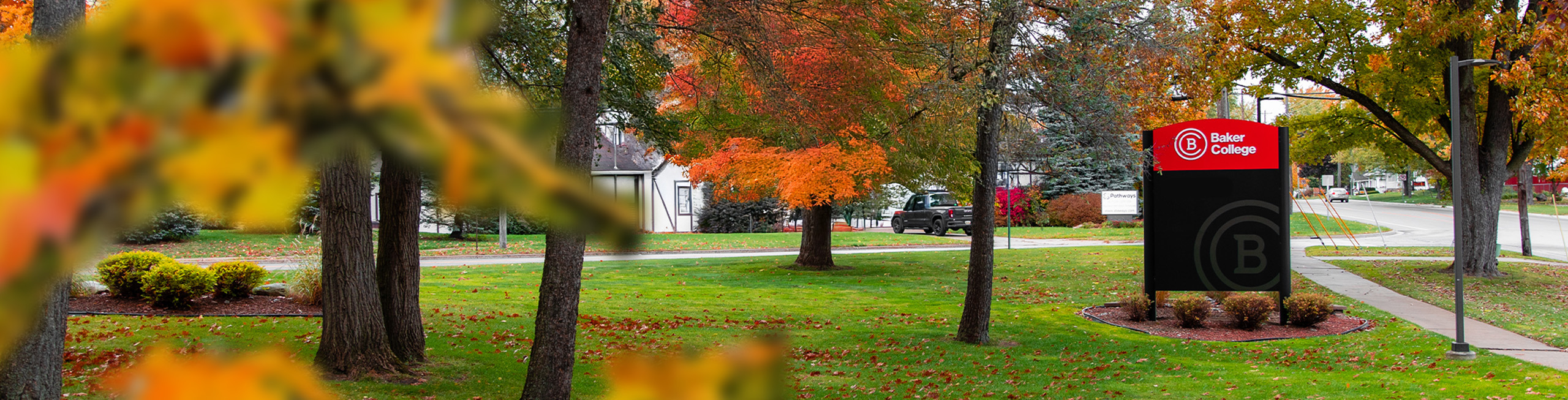A photo of the Baker Campus exterior with fallen leaves on the ground, as seen through trees. The right side is focused on a Baker College sign.