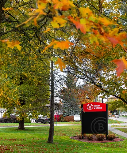 A photo of the Baker Campus exterior with fallen leaves on the ground, as seen through trees. The center is focused on a Baker College sign.