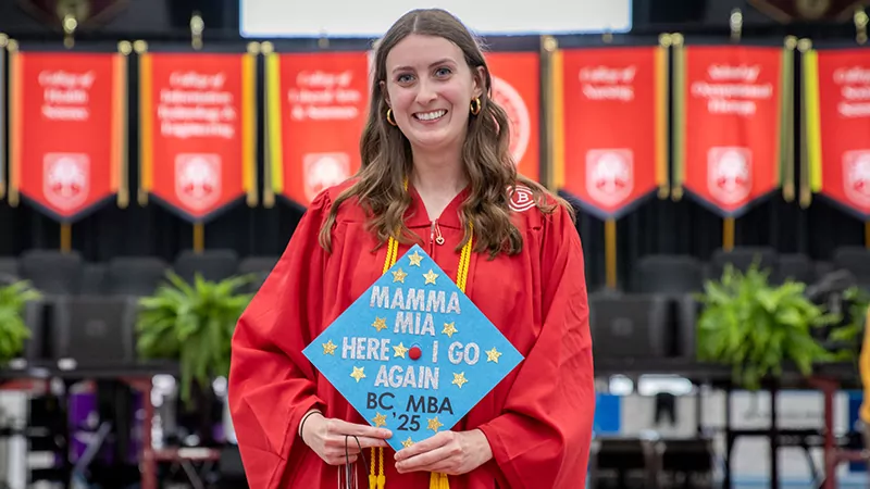 A woman holds her decorated graduation cap while smiling at Commencement