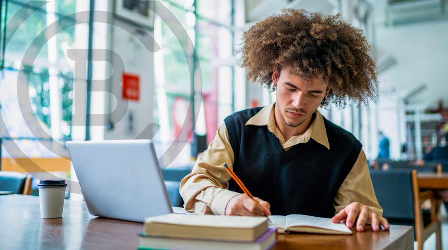 A student writes in a notebook next to a laptop and books.