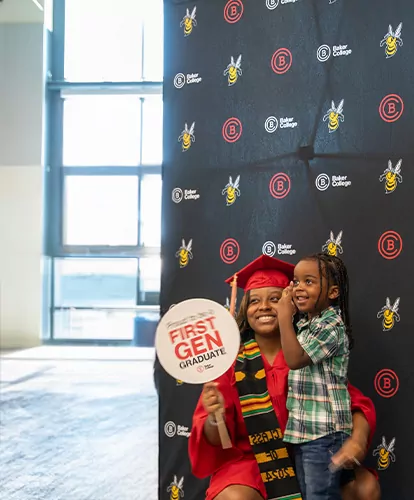 A graduate in a red cap and gown takes a photo with her son in front of a Baker College backdrop. The graduate holds a First Gen sign.