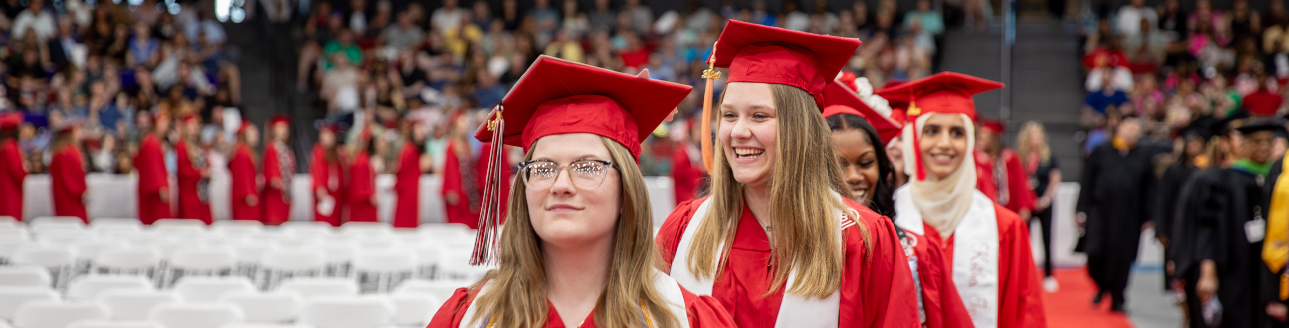 A line of smiling graduates walking in an auditorium full of spectators.