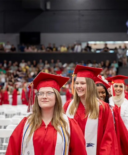 A line of smiling graduates walking in an auditorium full of spectators.