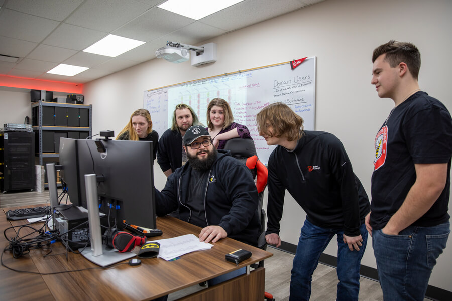 Cyber Defense Competition Team students work together on a challenge. One student sits at a desktop, while five others stand and contribute ideas.