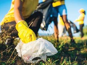 A close-up photo of a person's hands holding a trash bag. The person is wearing yellow gloves. Several others clean and hold trash bags behind them.