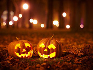 Two jack-o-lanterns lit from within with candles sitting on fallen leaves at night with streetlights in the background.