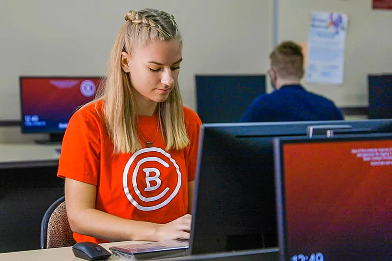 A student wearing a shirt with the Baker College logo types at a computer in a lab