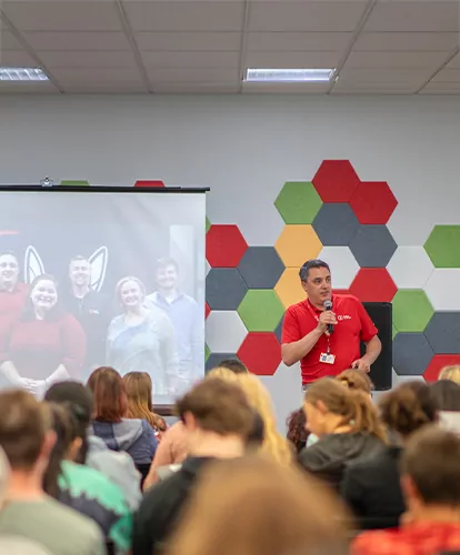 A Baker College staff member speaks to a crowd of prospective students. A slideshow can be seen on the hanging screen behind him.