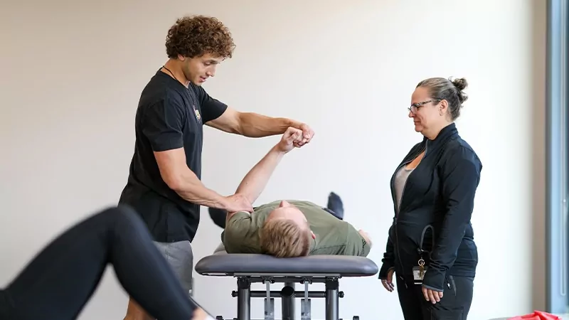 A standing student practices pysical therapy exercises with a student lying on a table as an instructor supervises.