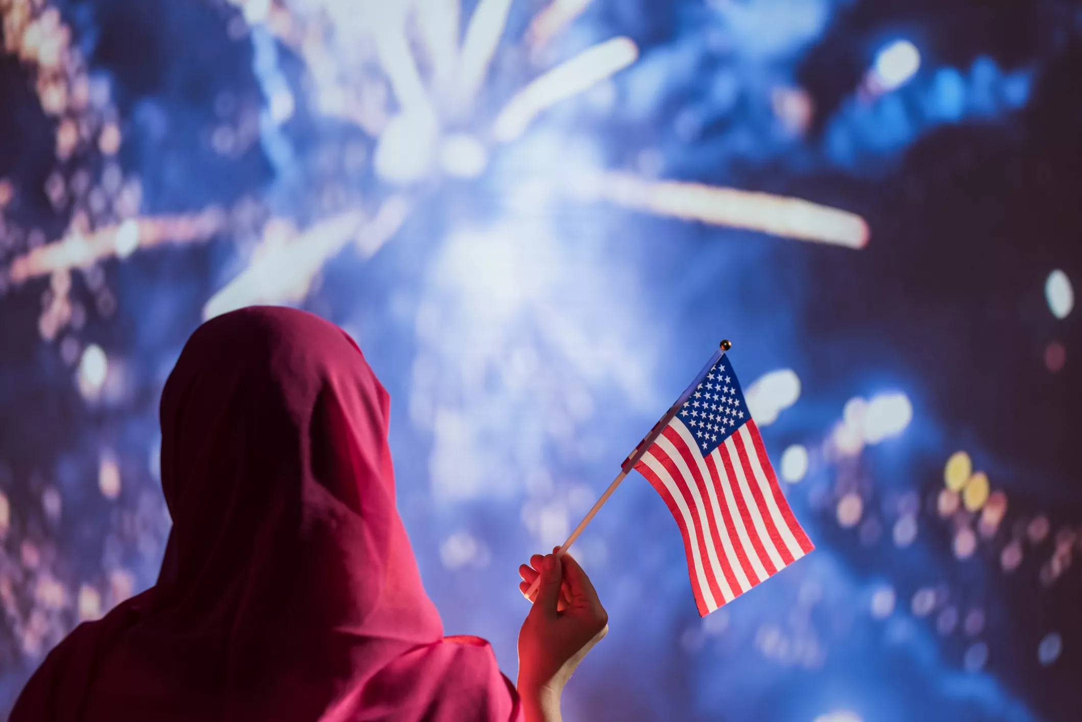 Arab American woman in a scarf holding American flag during fireworks at night.