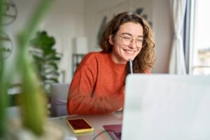 Happy young woman using laptop sitting at desk writing notes while studying online.