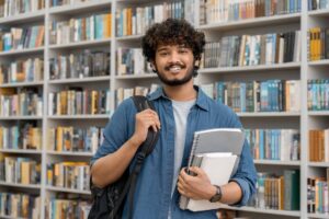 Portrait of cheerful male international Indian student with backpack, learning accessories standing near bookshelves at university library or book store during break between lessons.