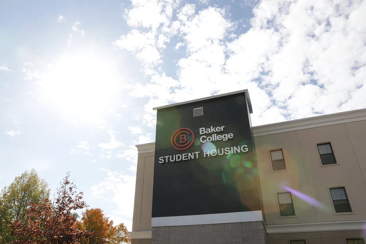 An exterior photo of the Student Housing sign with the Baker College logo against a bright blue sunny sky. A lens flare creates a reflection across the sign and housing building.