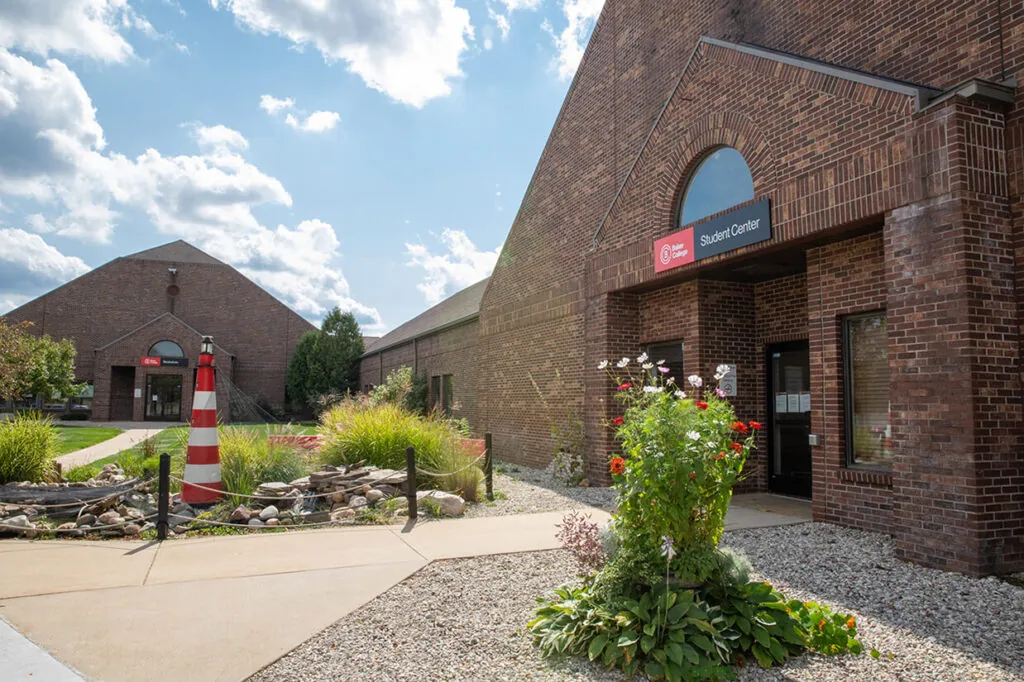 An exterior photo of the Student Center, a large brown brick building with a sign above the door, on the Cadillac campus next to a blue sunny sky with clouds.