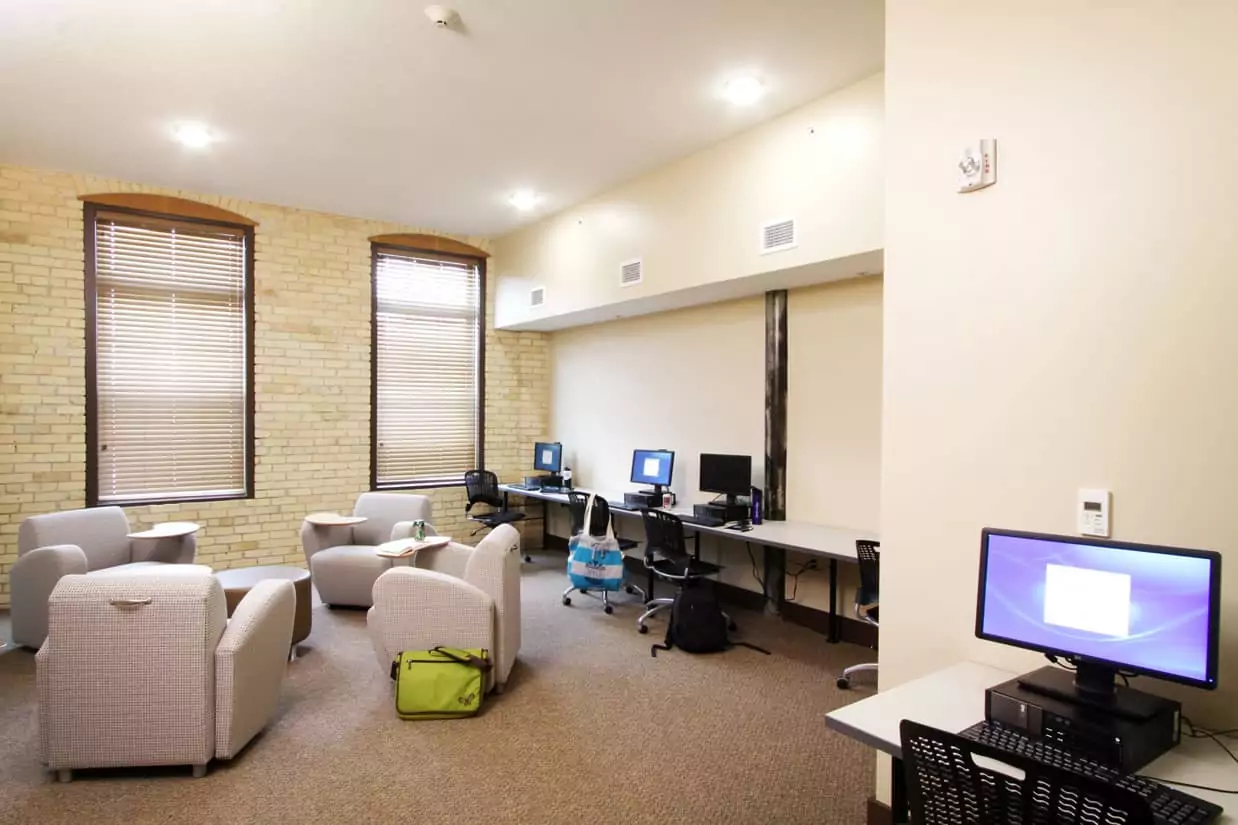 The interior of a study room at the Cadillac campus, a cozy space with one brick wall and a number of chairs with folding desk armrests in calming colors, along with several desktop workstations in a separate alcove.