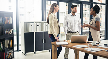 Three people in business attire stand and talk next to a desk with a laptop in an office.