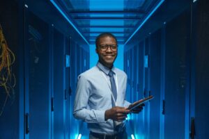 Smiling African American man standing by server cabinet while working with supercomputer in data center and holding tablet.