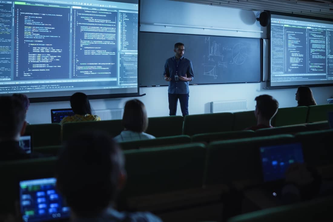 Young Male Teacher Giving a Data Science Lecture to Diverse Multiethnic Group of Female and Male Students in Dark College Room.