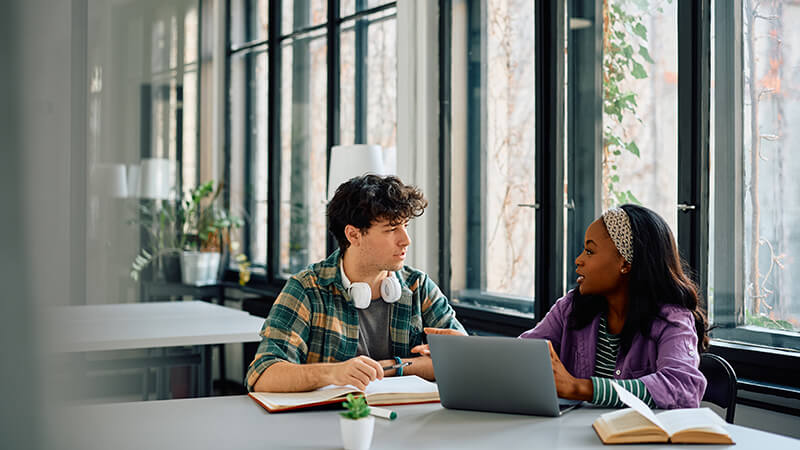 Two students chat over coursework at a table with a laptop and two books open in front of them.