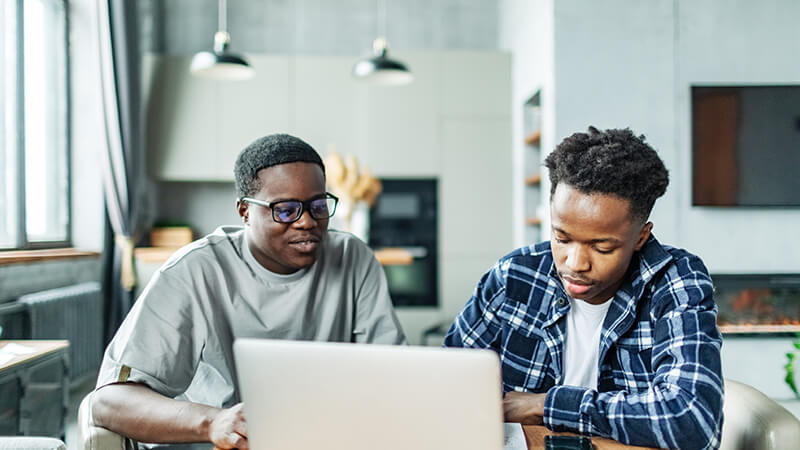 A man works through coursework as a friend sits nearby to help.