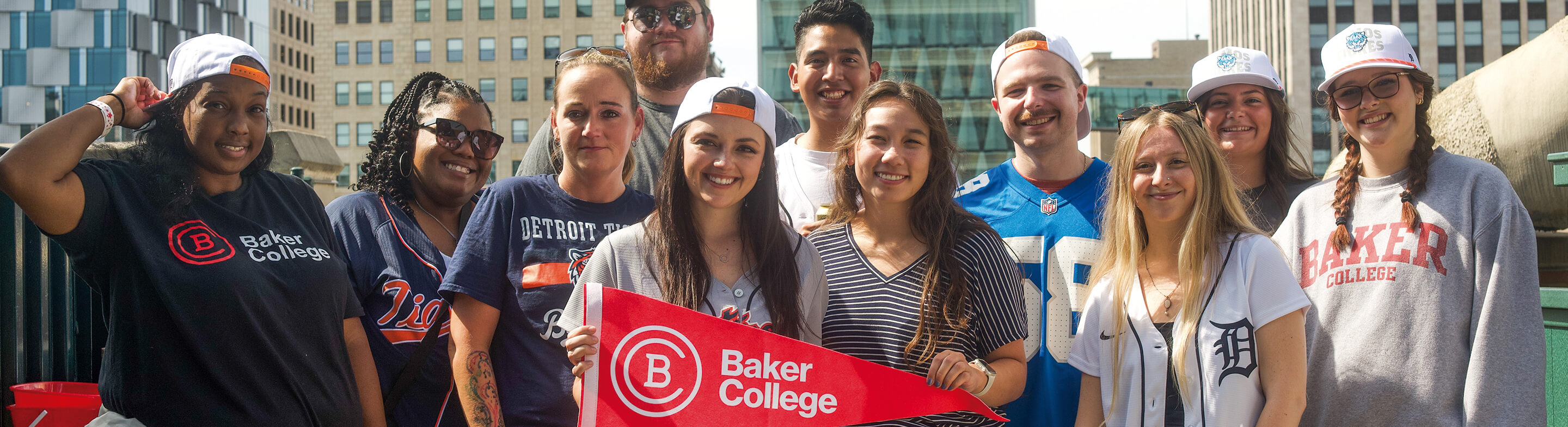 A group of students stand outside in front of tall city buildings while holding a Baker College pennant flag and smiling