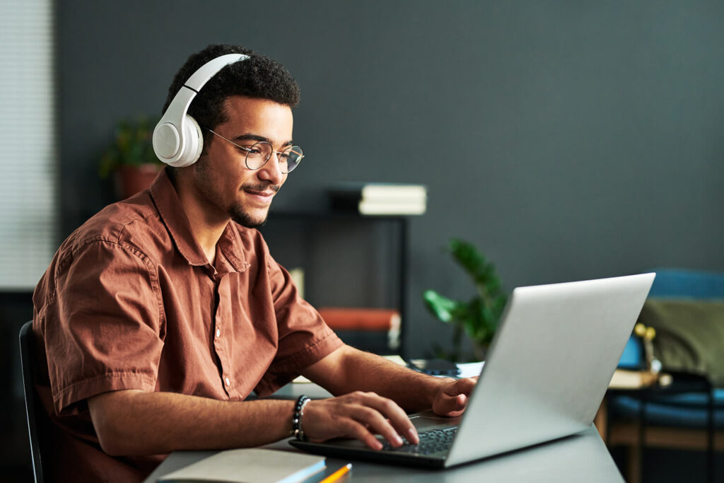 A young adult sits in front of a laptop with headphones on and a notebook nearby.