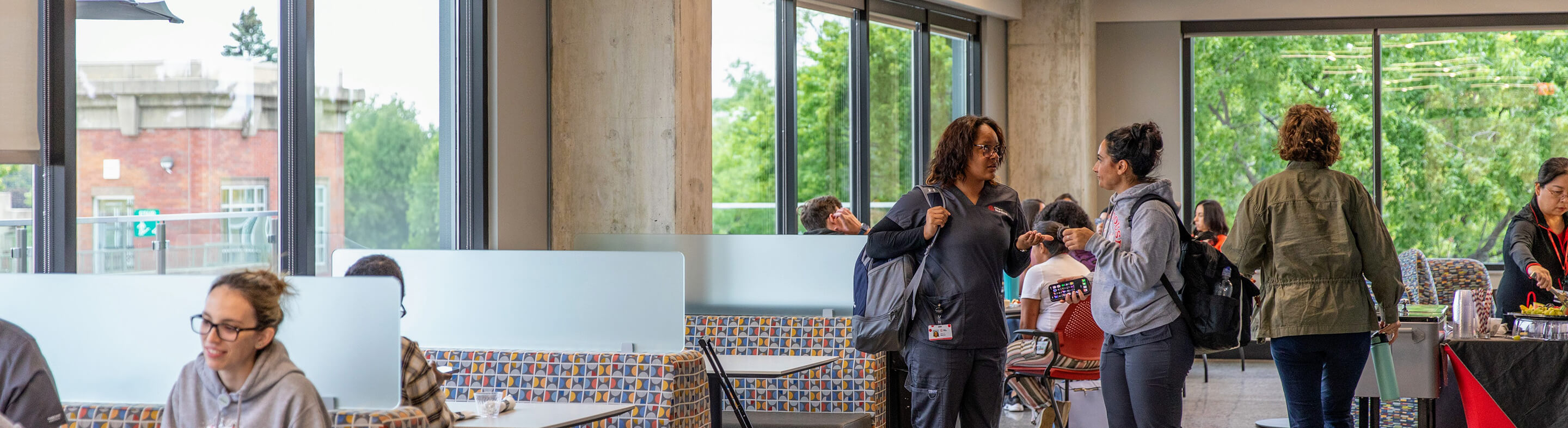 Students stand and chat on campus as other students study together while seated in booths.