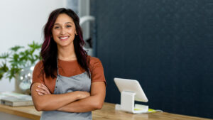 A woman wearing an apron stands proudly with her arms folded near a payment register.