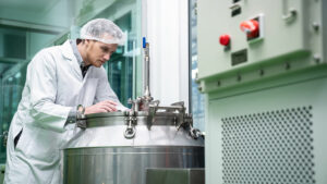 A lab worker in a lab coat and wearing a hairnet checks on a large steel boiler.