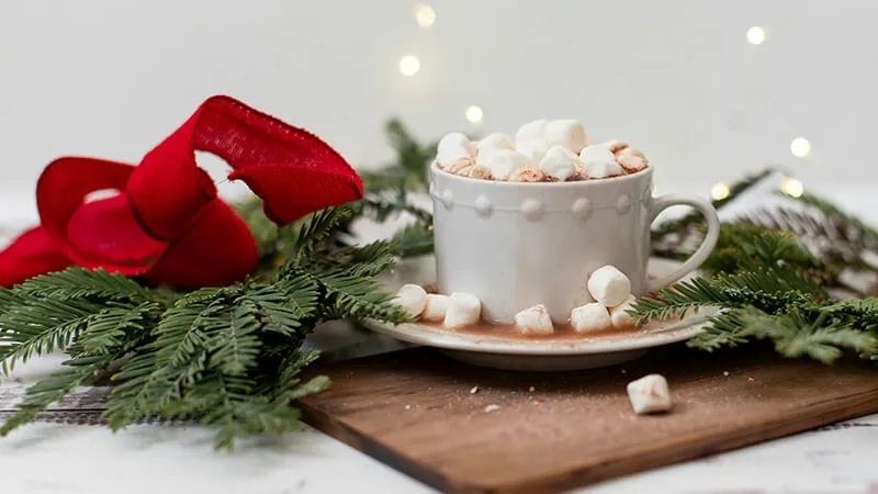 A mug of hot chocolate, topped with marshmallows, on a plate surrounded by pine branches, a red ribbon, and holiday lights.
