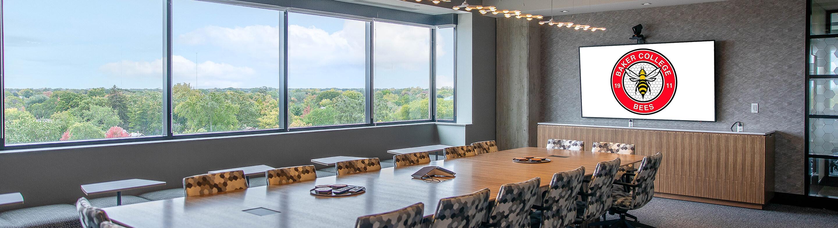 The Baker College Boardroom, a long wooden conference table surrounded by chairs. The Bees spirit mark is visible on a TV screen.