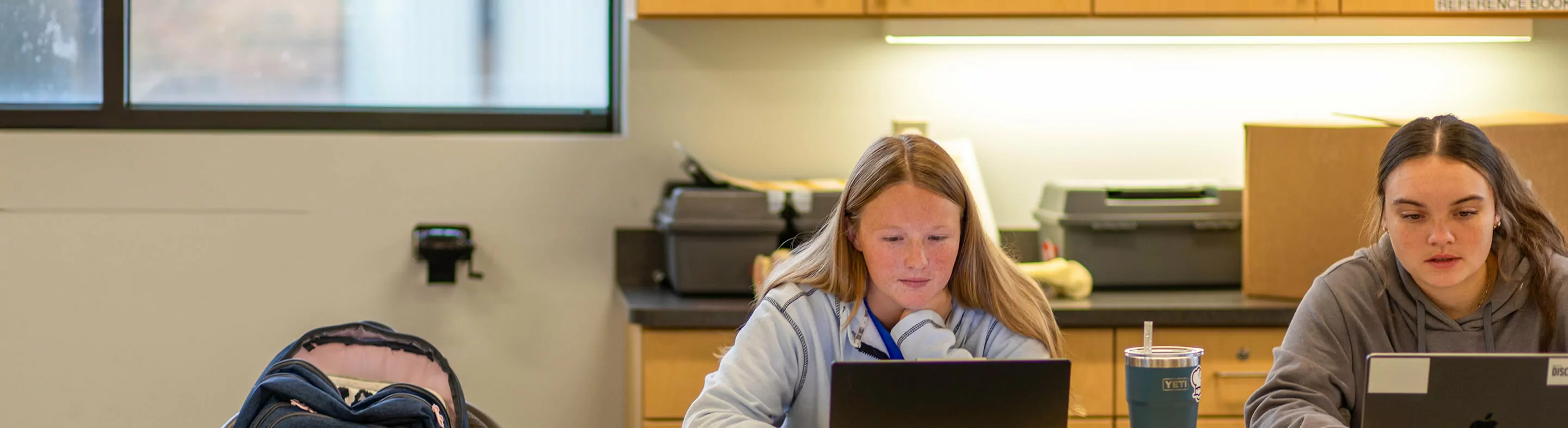 Two students type notes on laptops in a classroom.