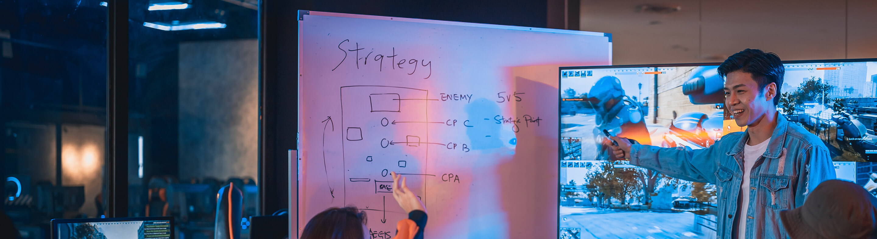 A student gestures to a large monitor showing a split-screen gaming session. A whiteboard showing game strategy sits nearby.