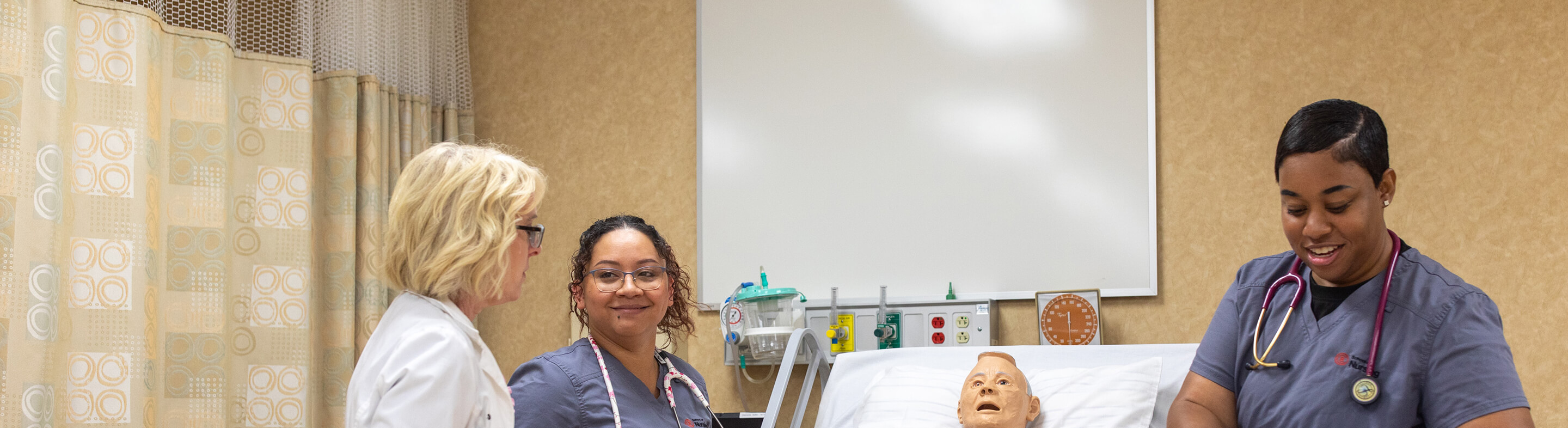 Two nursing students in scrubs work with a dummy while listening to their professor.