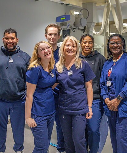 A group of students in scrubs smiles at the camera while standing near medical equipment.