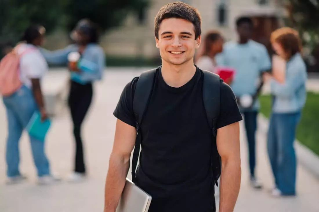 Young university student standing at college campus with laptop smiling at the camera.