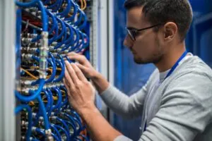 Side view portrait of young man connecting wires in server cabinet while working with supercomputer in data center.