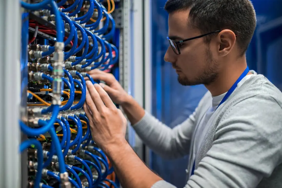 Side view portrait of young man connecting wires in server cabinet while working with supercomputer in data center.
