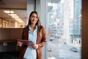 Young happy businesswoman using digital tablet while standing by the window in the office and looking at camera.
