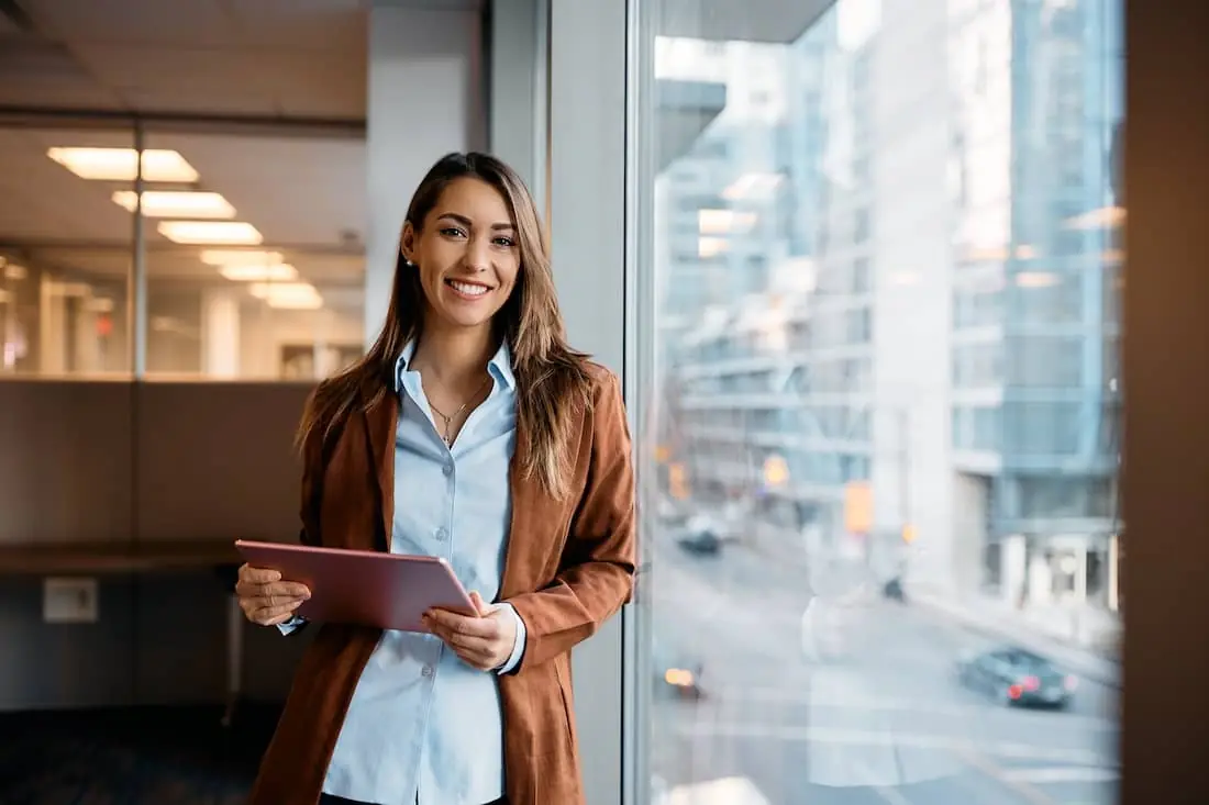 Young happy businesswoman using digital tablet while standing by the window in the office and looking at camera. 