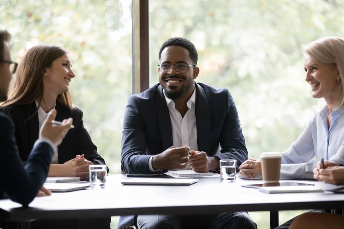 Five positive multi ethnic company staff gather together sit at boardroom desk talking planning future project.