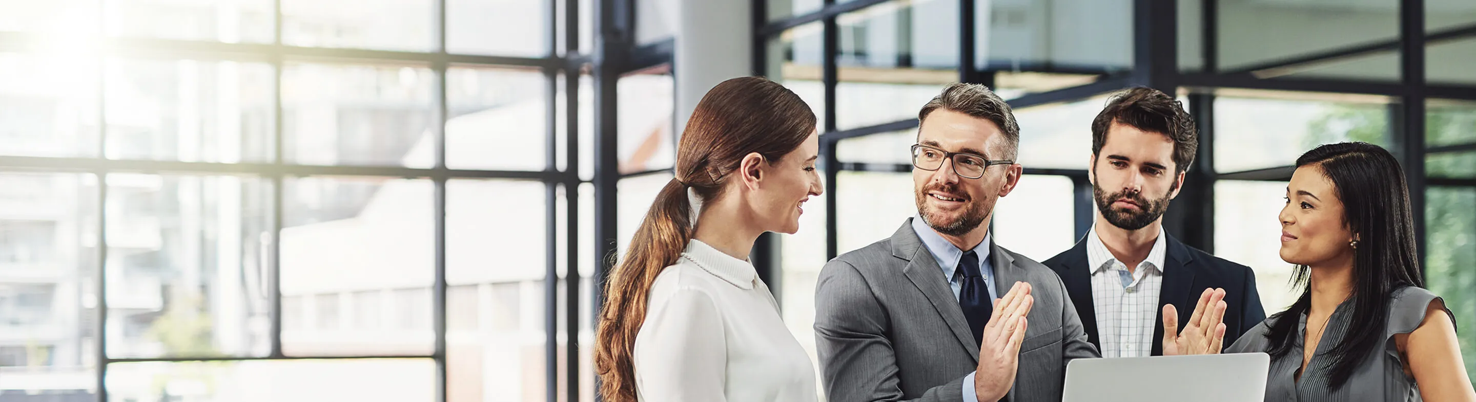 Four business professionals chat in a modern office with large class windows.