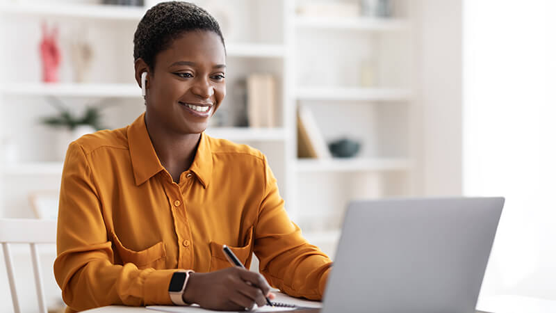 An African American woman smiles while holding a pen above a notebook. She's also watching her laptop screen and wearing ear buds.