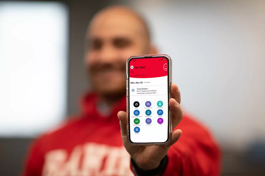 A student in a Baker College sweatshirt holds up a phone with the HiveConnect/Navigate Student app displayed on the screen.