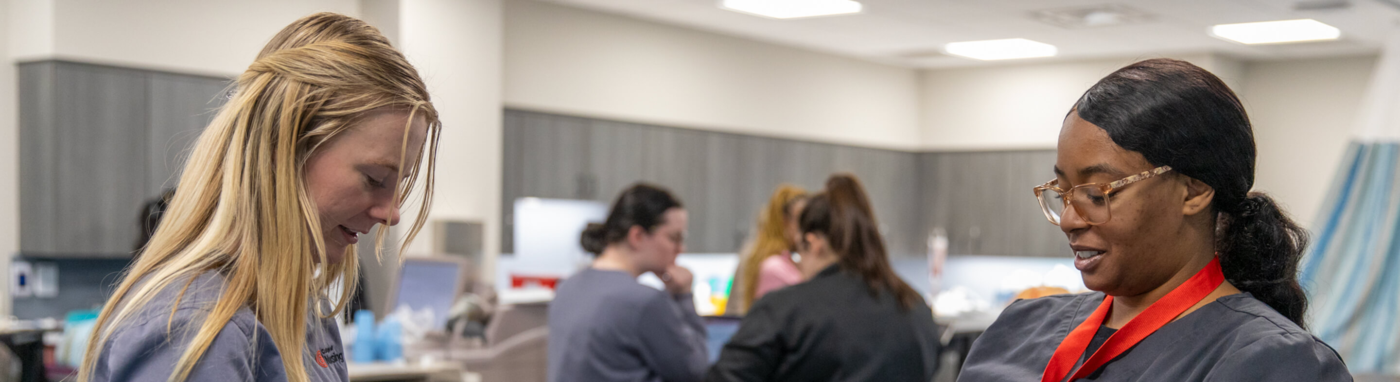 Two nursing students in scrubs work in a nursing lab while several others confer in the background.