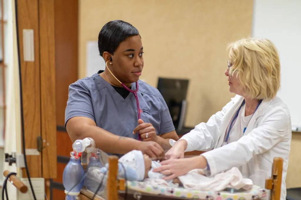 A nursing student wearing scrubs works with an instructor in a medical lab.
