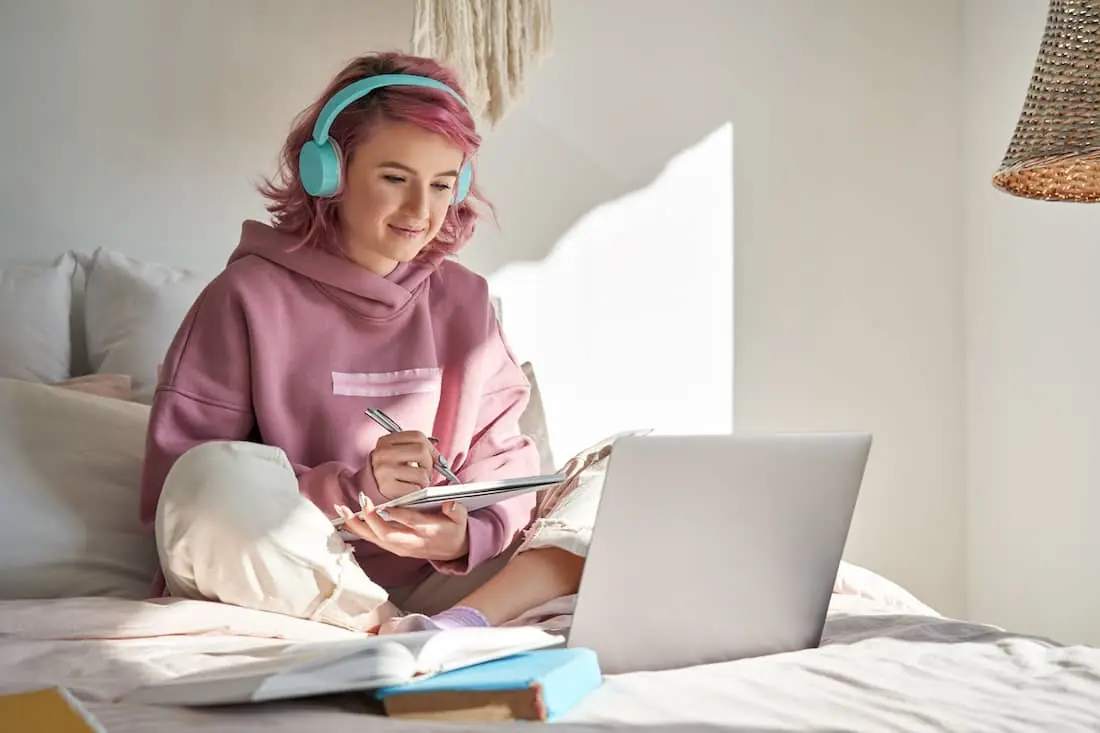 College student with pink hair wearing headphone writing notes and watching online webinar.