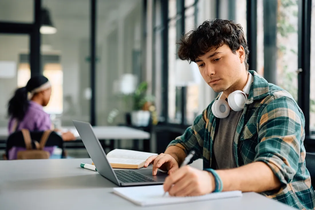 College student writing while using laptop and studying at a desk.