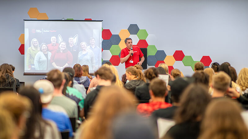 A speaker in a red Baker College polo holds a microphone and speaks to a crowd of seated students.