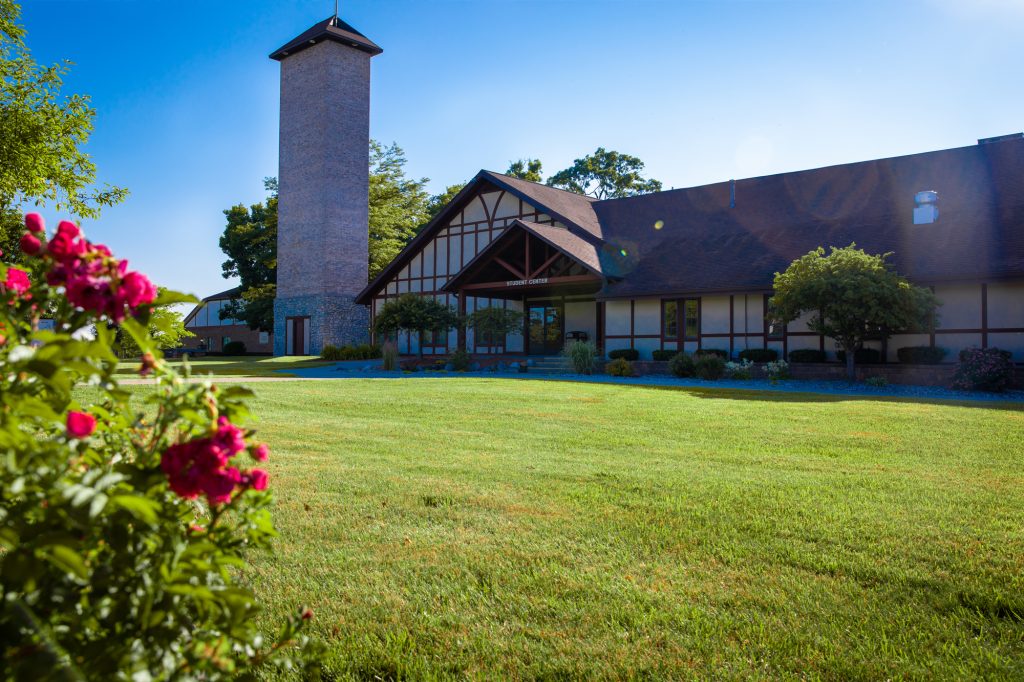 An exterior photo of the Owosso campus as seen next to a blue sky and a flowering plant.